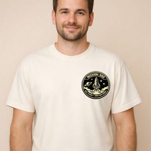 May include: A cream-colored short-sleeve t-shirt featuring a circular black and yellow graphic on the left chest. The design depicts a rocket launching into space with stars and a planet, including the text MISSION: DAD, EST. [YEAR], ORBIT, KID COUNT.