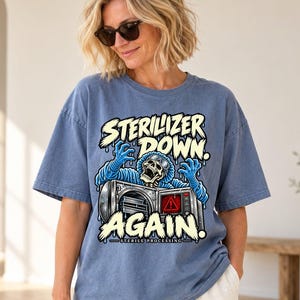May include: A blue short-sleeve t-shirt featuring a graphic of a skeleton in blue scrubs reaching out from an open autoclave machine. The text reads STERILIZER DOWN AGAIN and STERILE PROCESSING in a bold, light-colored, dripping font style.