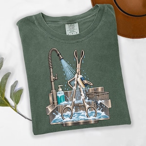 May include: A forest green t-shirt with a graphic print of a metallic surgical clamp standing in a stainless steel sink, scrubbing itself with a brush under a faucet stream. The shirt features a Comfort Colors brand label at the inner neckline.
