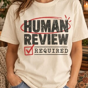 May include: A cream-colored short-sleeve t-shirt featuring the black distressed text "HUMAN REVIEW" circled in red, with a red checkbox containing a checkmark next to the black distressed text "REQUIRED" printed on the front of the garment.