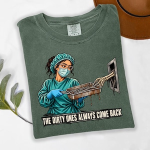 May include: A sage green t-shirt featuring a graphic of a medical professional in scrubs, a surgical cap, and a face mask holding a metal tray. A skeletal hand reaches from a wall opening toward the tray. The text reads, THE DIRTY ONES ALWAYS COME BACK.