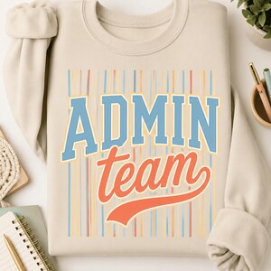 May include: A beige crewneck sweatshirt featuring a graphic print with vertical stripes in light blue, orange, and yellow. The text "ADMIN team" is printed in the center, with "ADMIN" in a blue block font and "team" in an orange script font.