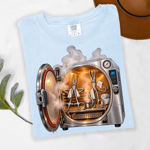 May include: A light blue t-shirt featuring a graphic of an open autoclave machine with two anthropomorphic surgical instruments inside. The instruments are sitting on a wooden bench with white towels, surrounded by steam, with a Comfort Colors label.
