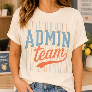 May include: A cream-colored short-sleeve t-shirt featuring a graphic print with vertical stripes in pastel colors. The text "ADMIN" appears in large blue block letters above the word "team" written in a cursive orange font with an underline.