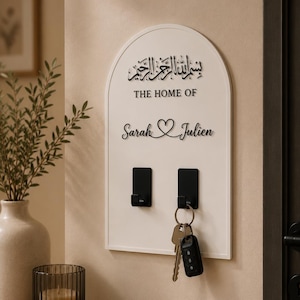Könnte beinhalten: Ein bogenförmiger, weißer Wand-Schlüsselhalter mit schwarzer arabischer Kalligrafie oben, dem Text "THE HOME OF" darunter und den Namen "Sarah" und "Julien", die durch ein Herzsymbol getrennt sind. Zwei schwarze Metallhaken sind angebracht.