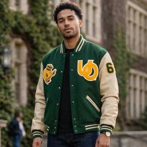 Può includere: Una giacca varsity con corpo in lana verde scuro e maniche color crema, chiusura con bottoni a pressione e polsini a coste a righe. Il petto presenta una toppa con papero giallo e una grande lettera gialla, con il numero 6 sulla manica.