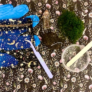 May include: A craft kit containing a pair of blue nitrile gloves, a dark blue velvet drawstring pouch with gold moon and star patterns, a small glass vial with a cork stopper, a clump of green moss, a dark soil disc, tweezers, and a plastic cup.