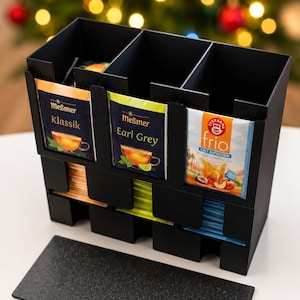 Könnte beinhalten: Ein schwarzer Teebeutel-Organizer mit drei vertikalen Fächern, die mit Teebeuteln gefüllt sind. Vorne sind Teebeutel von Meßmer Klassik, Meßmer Earl Grey und Teekanne Frio zu sehen. Ein separates rechteckiges schwarzes Bodenteil liegt flach davor.