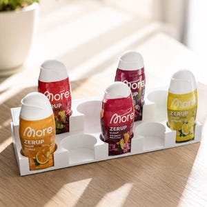 Könnte beinhalten: Ein weißer Kunststoff-Organizer mit vier Flaschen More Zerup zuckerfreiem Sirup. Die Flaschen haben orange, rote und gelbe Etiketten mit Fruchtillustrationen und weißen Verschlüssen. Das Tablett bietet sechs runde Fächer.