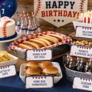 Könnte beinhalten: Ein Baseball-Party-Tisch mit Kuchen, Hotdogs, Hamburgern, Chips, Popcorn, Erdnüssen und Soda. Der Kuchen ist wie ein Baseball dekoriert. Schilder mit der Aufschrift "Happy Birthday", "Grand Slam Hot Dogs", "Batter Up Chips", "Ballpark Popcorn", "Peanuts & Cracker Jack", "Home Run Hamburgers" und "Slugger Soda".