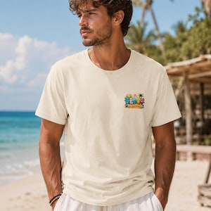 Può includere: T-shirt color crema con grafica colorata a tema spiaggia sul davanti. La grafica include il testo "Mr. Beach is Calling" con illustrazioni di cartoni animati di un sole, palme e personaggi da spiaggia. La maglietta viene indossata all'aperto.