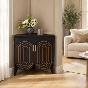 May include: A black corner cabinet featuring two doors with gold-toned concentric oval patterns and vertical gold handles. The top surface holds a dark gray ribbed vase filled with white and yellow flowers alongside a small glass candle holder.