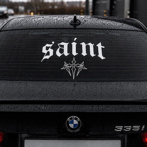 Puede incluir: Una calcomanía de vinilo blanco aplicada en la ventana trasera de un coche negro. La calcomanía presenta la palabra saint en una fuente de estilo gótico, colocada sobre un gráfico de cruz estilizada. La ventana tiene gotas de lluvia.