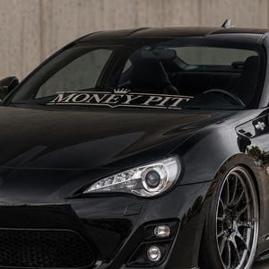 Puede incluir: Un coche deportivo Toyota negro con una pegatina blanca en el parabrisas que dice MONEY PIT con un pequeño icono de corona sobre el texto. El vehículo tiene un capó liso, faros claros y llantas oscuras de radios múltiples con acabado metálico.