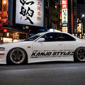 Peut inclure: Un coupé blanc à deux portes garé dans une rue urbaine la nuit. La voiture est équipée de jantes sombres à rayons multiples avec des bords polis et d'autocollants noirs sur le côté indiquant KANJO STYLE, OSAKA STREET DIVISION et des caractères japonais.
