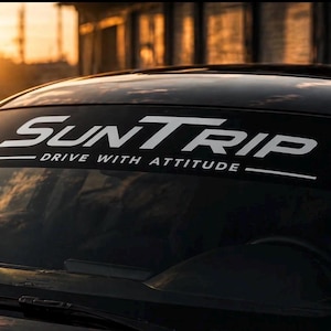 Puede incluir: Una franja parasol negra aplicada en la parte superior del parabrisas de un coche. El adhesivo presenta el texto blanco SUNTRIP en una fuente estilizada en negrita, con la frase DRIVE WITH ATTITUDE impresa en letras blancas más pequeñas.
