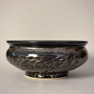 May include: A wide, low-profile ceramic bowl with a glossy black glaze featuring a textured, metallic silver-toned finish around the midsection. The bowl has a smooth, rounded rim and a distinct circular base with small drainage holes visible.