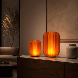 May include: Two cylindrical table lamps with vertical ribbed glass textures in a warm orange hue, emitting a soft internal glow. The lamps are positioned on a surface alongside a plain, light gray ceramic vase with a rounded body and narrow neck.