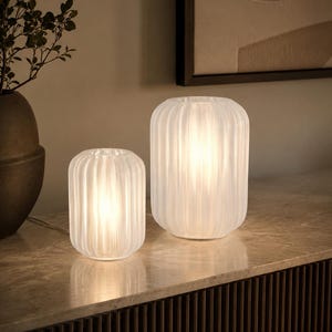 May include: Two cylindrical table lamps made of translucent, ribbed white glass. Each lamp features a vertical fluted texture that diffuses the internal light, creating a soft glow. The lamps are of different heights, with one smaller and one larger.