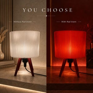 May include: A table lamp with a cylindrical, vertically ribbed shade and three dark wooden legs. The text YOU CHOOSE appears above, with Without Red Insert on the left showing white light and With Red Insert on the right showing red light.