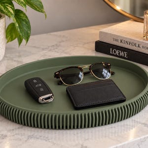 May include: A round olive green tray with a ribbed exterior edge. Resting on the tray are a black car key fob with silver buttons, a pair of tortoiseshell-rimmed sunglasses with gold-tone accents, and a small black leather card holder wallet.