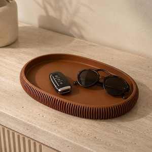 May include: An oval-shaped, terracotta-colored tray with a ribbed, textured exterior edge. The tray holds a black car key fob and a pair of dark-rimmed sunglasses with brown tortoiseshell-patterned frames resting on its smooth, flat interior surface.