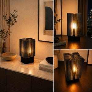 May include: A table lamp featuring a square base and a series of vertical, translucent gray glass panels arranged in a cross-like formation. The lamp emits a warm, soft glow from within, highlighting the ribbed texture of the glass structure.