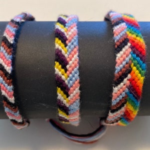 May include: Three colourful friendship bracelets with a chevron pattern. The bracelets are made with different coloured threads, including pink, blue, yellow, red, orange, brown, black and white.