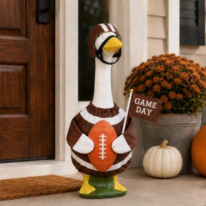 May include: A white goose statue wearing a brown and white striped jersey, a brown football helmet, and yellow feet. The goose holds an orange football and a small brown flag with the white text GAME DAY printed on it, standing on a green base.