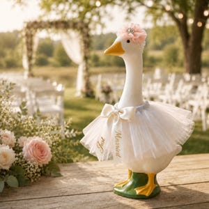 May include: A white goose figurine wearing a pale pink floral headpiece with a central crystal accent and a white tulle skirt featuring a satin bow with the text Just Married printed in gold script on the ribbon tails, standing on a green base.