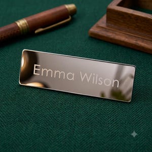 May include: A rectangular, polished silver-toned metal nameplate featuring the engraved name Emma Wilson in a clean, sans-serif font. The surface is highly reflective, showing subtle light highlights across its smooth, metallic finish.