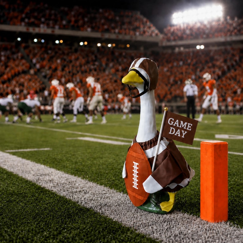 May include: A white goose figurine wearing a brown and white football helmet and a brown and white jersey with a football graphic on the chest. The goose holds a small brown flag with the white text GAME DAY, standing next to an orange pylon.