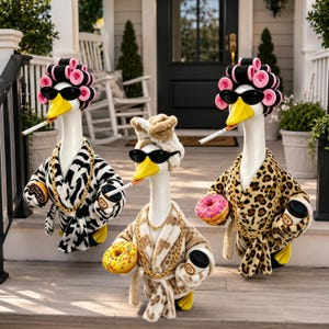 May include: Three white goose figurines wearing plush robes with animal prints, pink hair rollers, and black sunglasses. Each goose holds a doughnut and a coffee cup, with a cigarette in its beak. They are adorned with gold-tone chain necklaces.