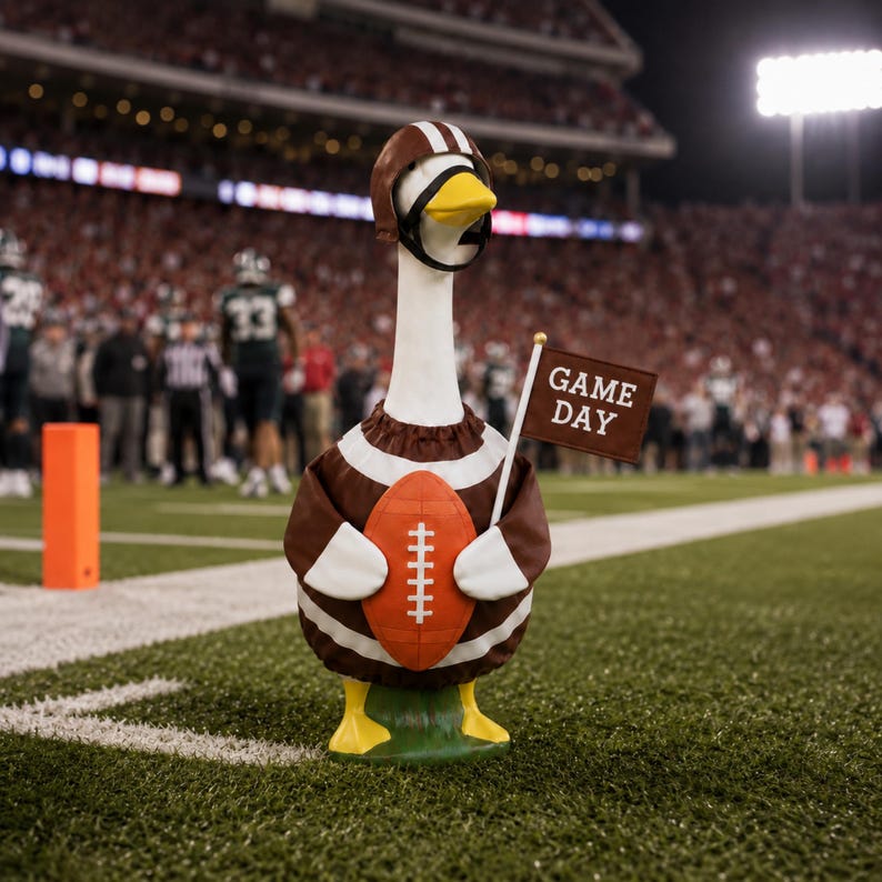 May include: A decorative goose figurine wearing a brown and white football jersey and a brown helmet with white stripes. The goose holds an orange football and a small brown flag with the white text GAME DAY printed on it, standing on grass.