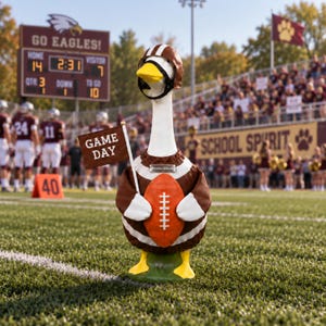 May include: A white goose figurine wearing a brown and white football jersey, a brown helmet with a black face mask, and yellow feet. The goose holds an orange football and a brown flag with the white text GAME DAY. A silver nameplate reads Emma Wilson.