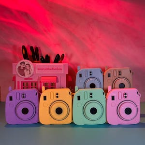 May include: A collection of colorful, square-shaped camera cases in shades of purple, yellow, green, and pink. Each case has a circular lens and a small viewfinder. The cases are arranged in a row, with a pink building-shaped pen holder in the background.