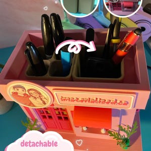 Peut inclure: Un organisateur de bureau rose en forme de petit magasin avec un portrait circulaire de deux personnes et du texte sur le devant. Il comporte plusieurs compartiments pour stylos et outils, dont deux organisateurs beiges d&eacute;tachables &agrave; l'int&eacute;rieur.