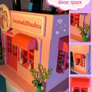 Peut inclure: Un organisateur de bureau rose en forme de boutique miniature avec des auvents orange, des cadres de fen&ecirc;tre rouges et une enseigne indiquant materialized.ca. Il comprend des petits vases blancs avec des plantes et des emplacements pour stylos.