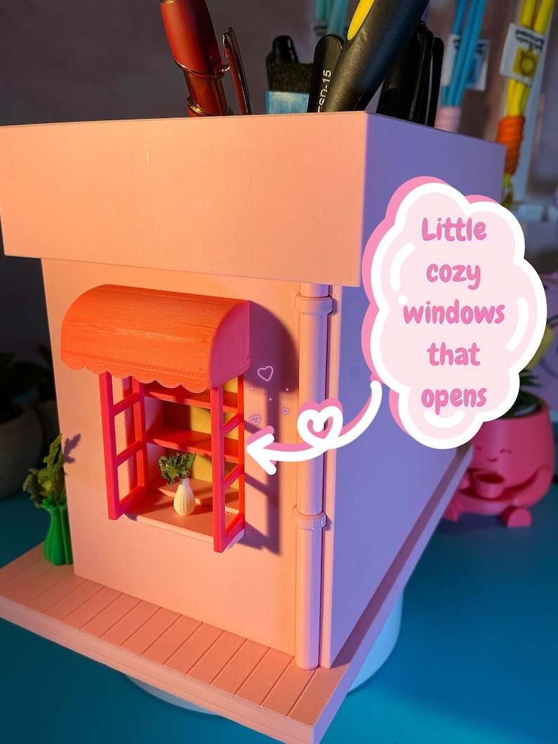 Peut inclure: Un organisateur de bureau rose clair en forme de b&acirc;timent avec un auvent orange vif au-dessus d'une fen&ecirc;tre. Le cadre de la fen&ecirc;tre est rouge et contient un petit vase blanc avec une plante verte. Une bulle rose avec le texte Little cozy windows that opens pointe vers la fen&ecirc;tre avec une fl&egrave;che blanche.