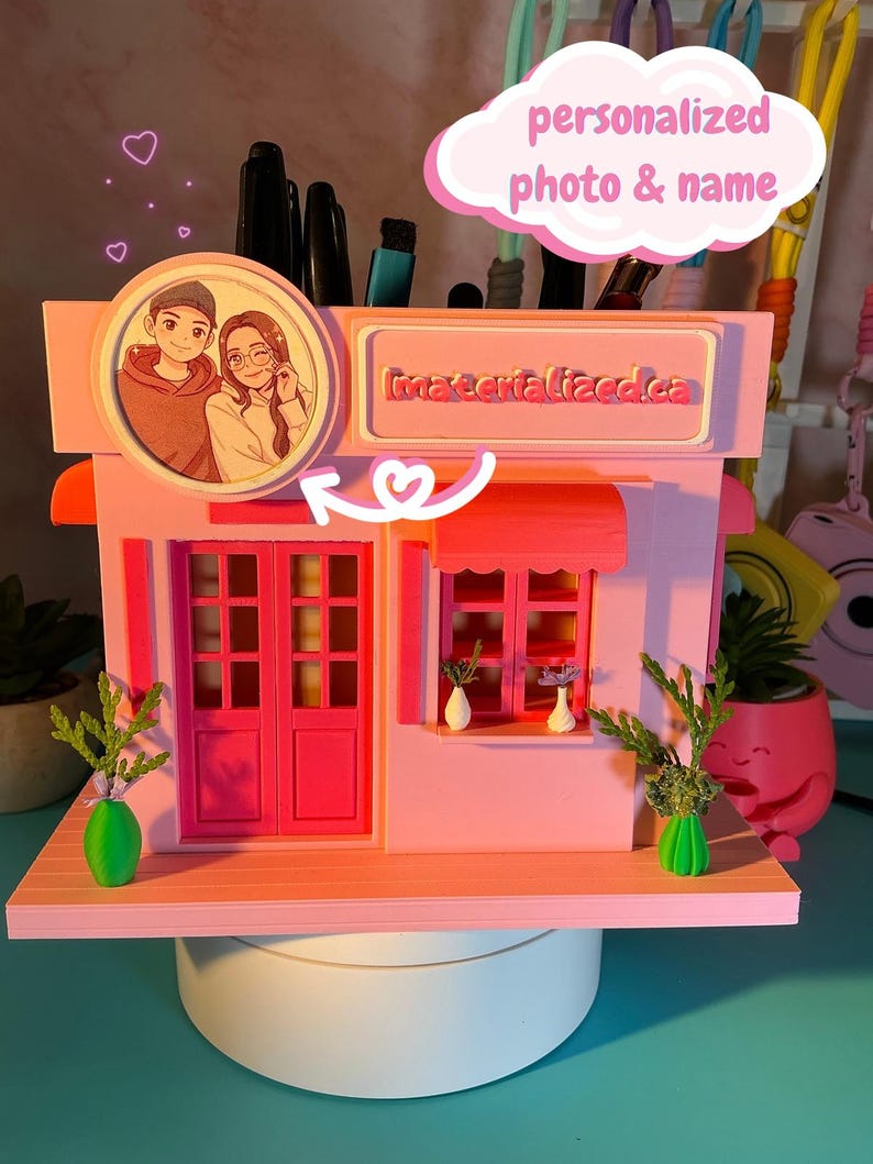 Peut inclure: Un organisateur de bureau rose en forme de petite devanture de magasin avec une illustration circulaire de deux personnes, une enseigne materialized.ca, une double porte, une fen&ecirc;tre avec deux petits vases blancs et des plantes vertes.
