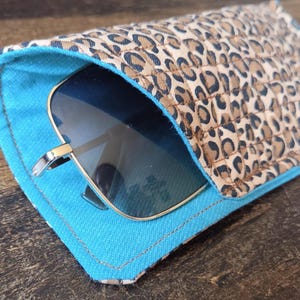 Puede incluir: Un estuche para gafas de tela acolchada con un exterior de estampado de leopardo en tonos beige y negro y un forro interior de color azul turquesa brillante. Unas gafas de sol oscuras con montura de metal plateado están dentro del estuche.