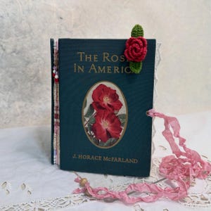 May include: A dark teal hardback book titled The Rose in America by J. Horace McFarland, featuring a central oval illustration of two red roses, a green crocheted rose bookmark, decorative page edges, and a long, crinkled pink ribbon tie.