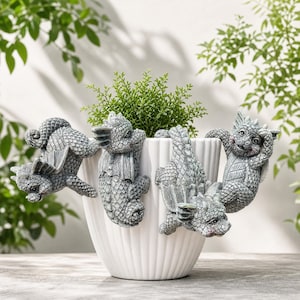 May include: A white, vertically ribbed ceramic plant pot featuring four grey, textured dragon figurines attached to the rim. Each dragon is depicted in a different pose, with detailed scales, small wings, and distinct facial expressions.