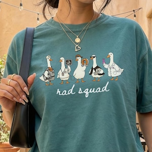 Puede incluir: Una camiseta de manga corta color verde azulado con un gráfico de cinco patos blancos vestidos con varios atuendos profesionales y el texto "rad squad" impreso en cursiva blanca debajo de la ilustración. La camiseta tiene cuello redondo.