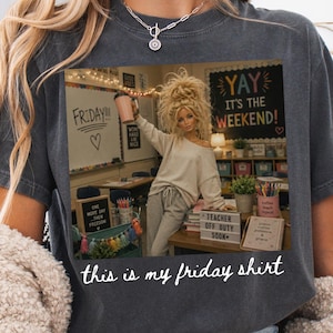 May include: A charcoal gray t-shirt featuring a graphic print of a doll in a classroom setting with the text "YAY IT'S THE WEEKEND!" and "FRIDAY!!!" on a whiteboard, with the white cursive text "this is my friday shirt" printed below the image.