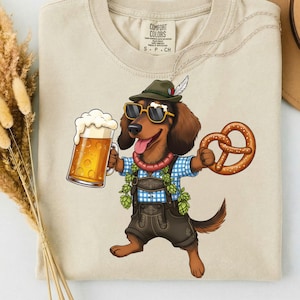 Könnte beinhalten: Ein beiges T-Shirt mit einer Grafik eines braunen Dackels, der einen grünen Hut, eine Sonnenbrille und eine Lederhose trägt. Der Hund hält einen großen Bierkrug und eine Brezel. Das T-Shirt hat ein Comfort Colors Etikett am Halsausschnitt.