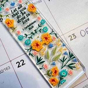 Peut inclure: Un marque-page rectangulaire transparent orné d'illustrations florales colorées en jaune, bleu sarcelle, rose et vert. Le texte Let all that you do be done in love, 1 Corinthians 16:14 est imprimé en noir parmi les motifs de fleurs peintes.