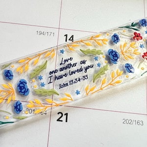 Peut inclure: Un marque-page rectangulaire en acrylique transparent orné d'un motif floral coloré avec des fleurs bleues, des touches rouges et des branches feuillues jaunes. Le texte indique : Love one another as I have loved you, John 13:34-35.