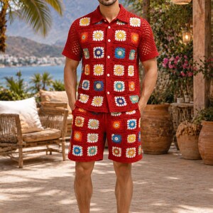 Op de afbeelding: Een rode gehaakte set bestaande uit een overhemd met korte mouwen en knopen en een bijpassende korte broek, voorzien van een kleurrijk granny square patroon met witte, gele en blauwe bloemmotieven, gecombineerd met effen witte sneakers.