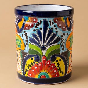 May include: A cylindrical ceramic container with a vibrant floral design. The container has a dark blue rim and base, with a white interior. The exterior is adorned with colorful flowers in red, yellow, orange, and green against a light blue background.